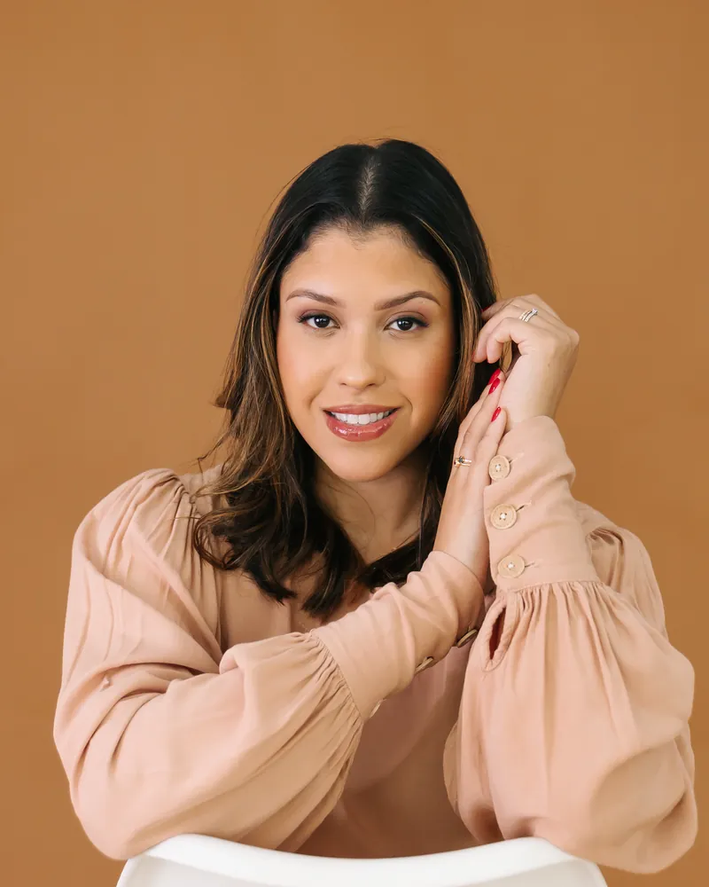 Emanuelle Mendes, psicóloga clínica, sorrindo com a mão no rosto, diante de um fundo marrom