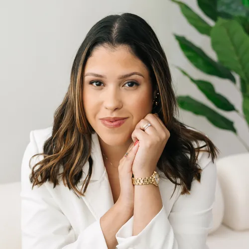 Emanuelle Mendes de blazer branco, sorrindo com o rosto apoiado nas mãos