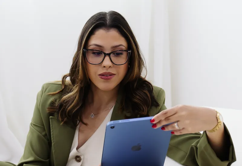 Emanuelle Mendes de óculos e blazer verde, sentada, olhando para o tablet