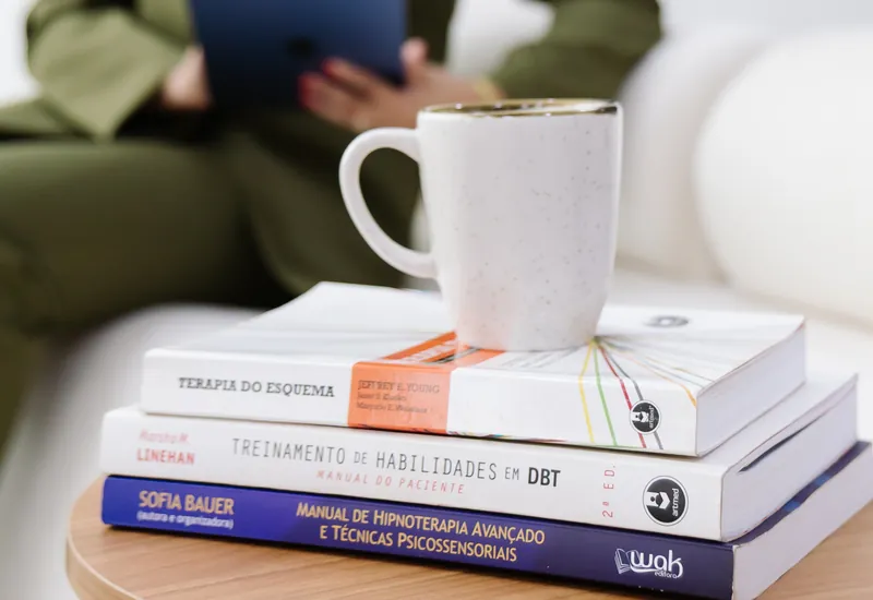 Livros de psicologia empilhados na mesa, com Emanuelle Mendes ao fundo